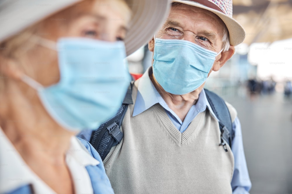 Senior gray-haired male tourist in a disposable face mask looking fixedly at his thoughtful wife