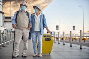Full-sized portrait of a man with a walking cane and his wife with a trolley suitcase standing outside