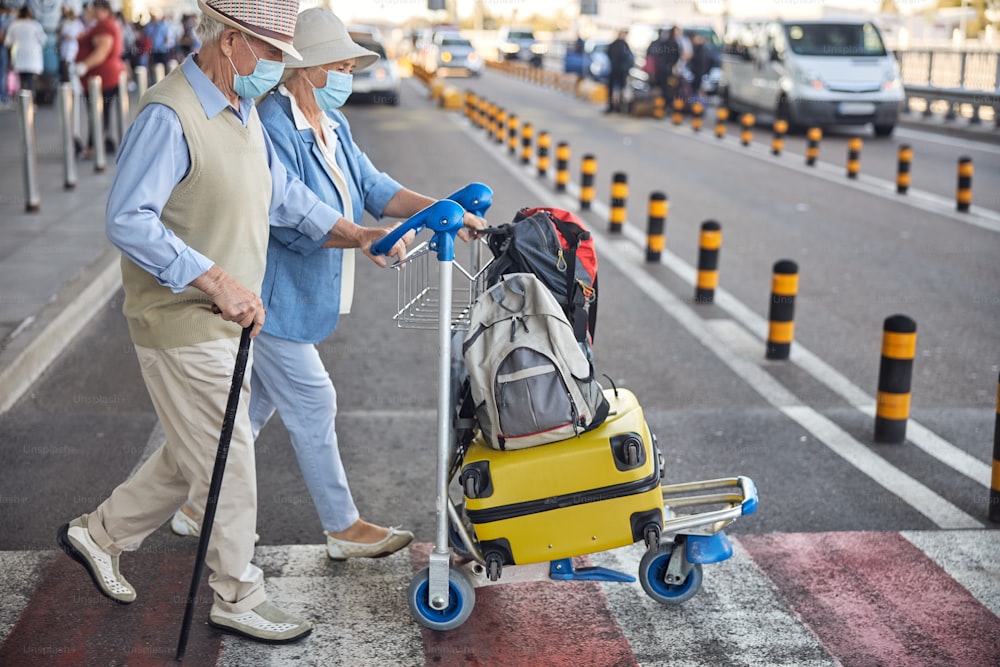Side view of an aged married couple in disposable face masks wheeling their baggage trolley