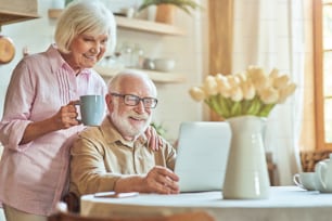 Mujer mayor sonriente y hermosa sosteniendo una taza de café y de pie cerca de su esposo mientras mira el video en la computadora. Concepto de estilo de vida doméstico