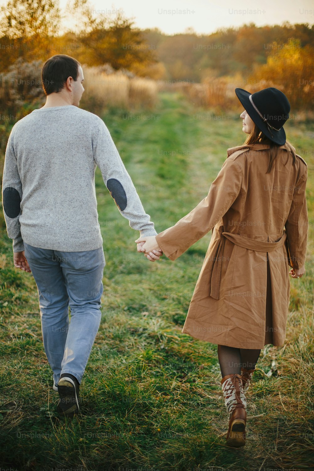 暖かい夕焼けの光の中で秋の牧草地を手をつないで歩く幸せなスタイリッシュなカップル。ロマンチックな官能的な瞬間。秋の野原でリラックスする若いファッショナブルな女性と男性