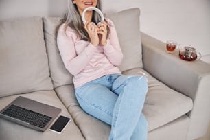 Senhora alegre segurando fones de ouvido sem fio e sorrindo enquanto descansa no sofá confortável com notebook e celular