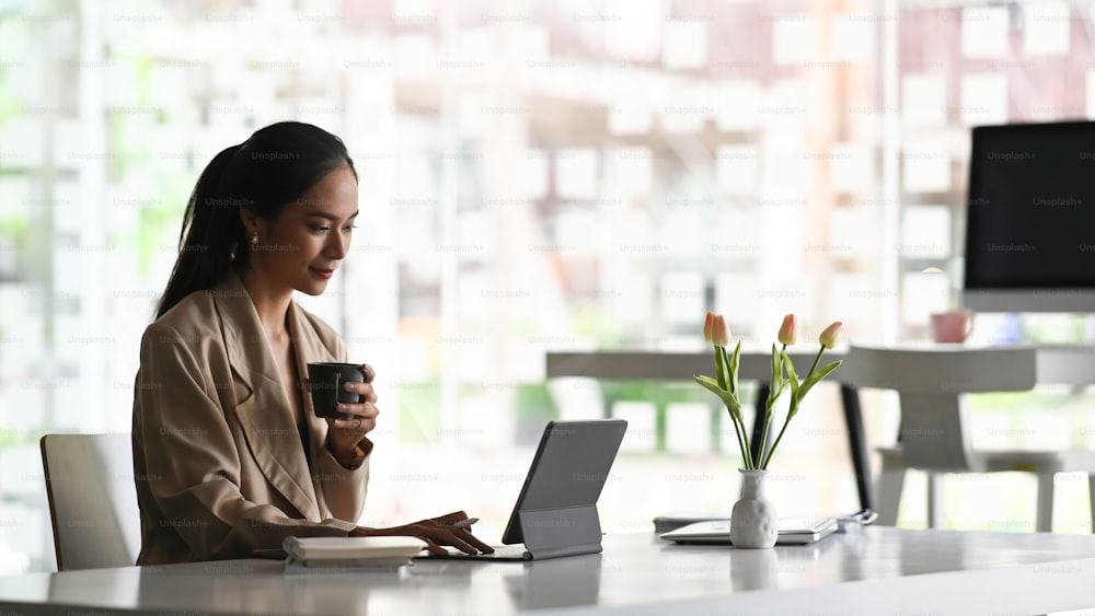 A young attractive woman working in new project with tablet while drinking coffee in modern office.