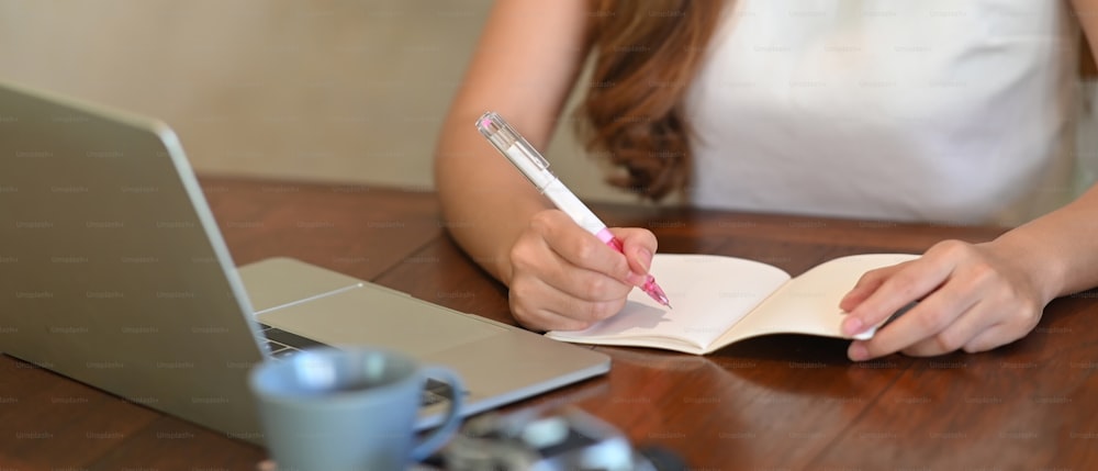 Cropped shot of young businesswoman sitting in front of laptop and taking notes in notebook.