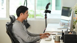 Sideview of photographer editing photos after a photo shoot on desktop with camera and lens placed on table.