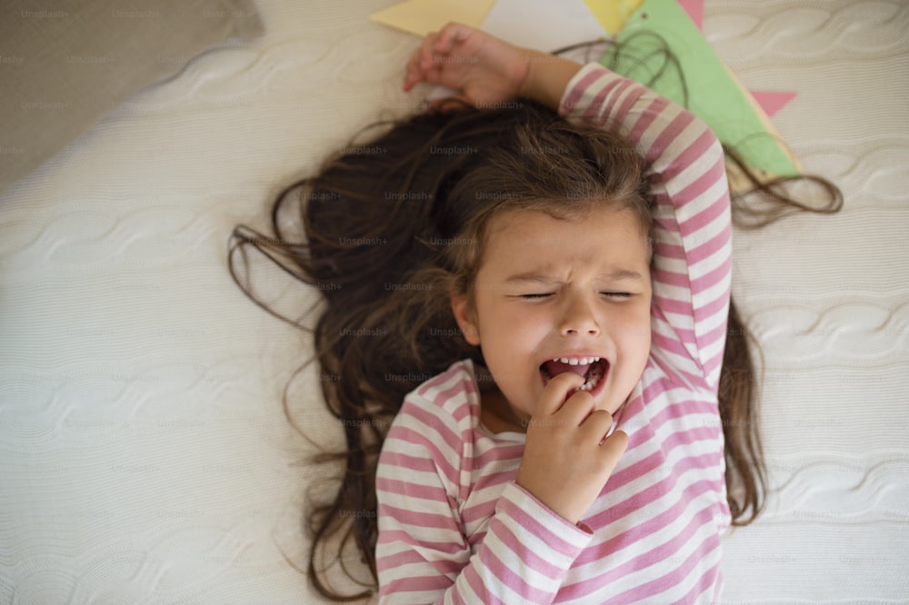 Top view portrait of unhappy small girl lying on bed indoors, crying.