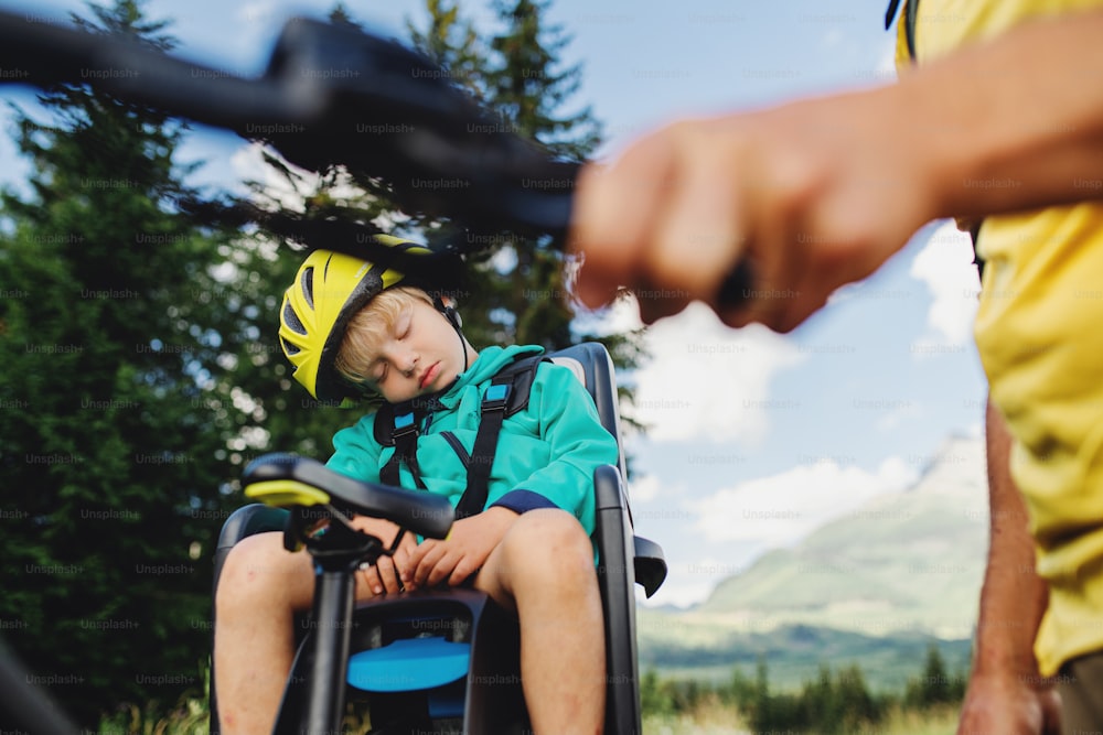 Unrecognizable father with tired sleeping small son cycling outdoors in summer nature.
