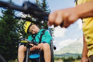 Unrecognizable father with tired sleeping small son cycling outdoors in summer nature.
