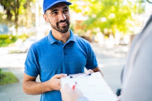 Portrait of a delivery man carrying packages while customer putting signature in clipboard. Delivery and shipping concept.