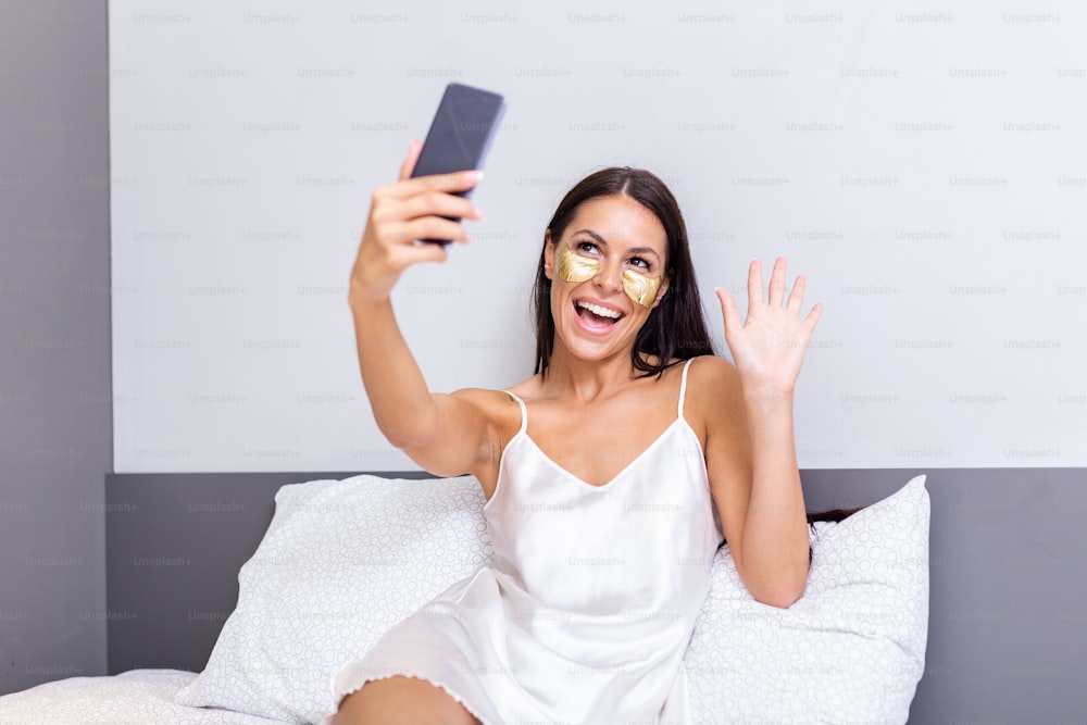 Sexy beautiful woman with Gold Cosmetics mask, Eye Patches with a smartphone in her bed. Beautiful young woman in silk nightgown reading text message on her mobile phone and smiling.