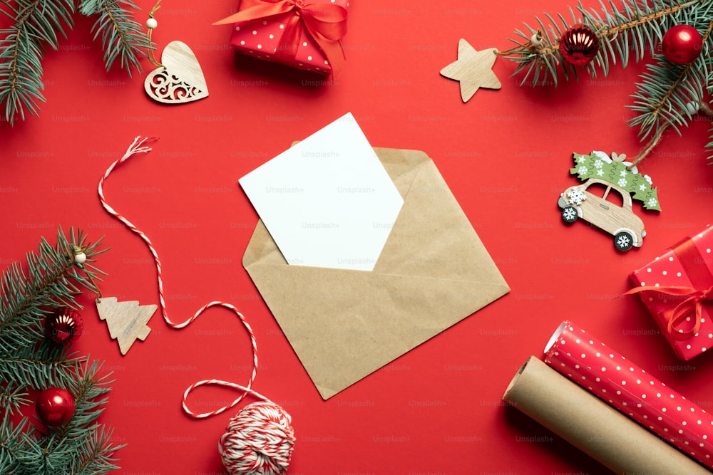 Craft paper envelope with empty card inside and Christmas decorations, gifts, fir tree branches on red background. Christmas letter concept.