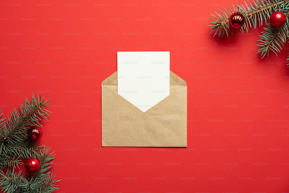 クリスマスの手紙のコンセプト。ビンテージクラフト紙の封筒に、中に空白の白いカードと赤い背景にモミの木の枝。