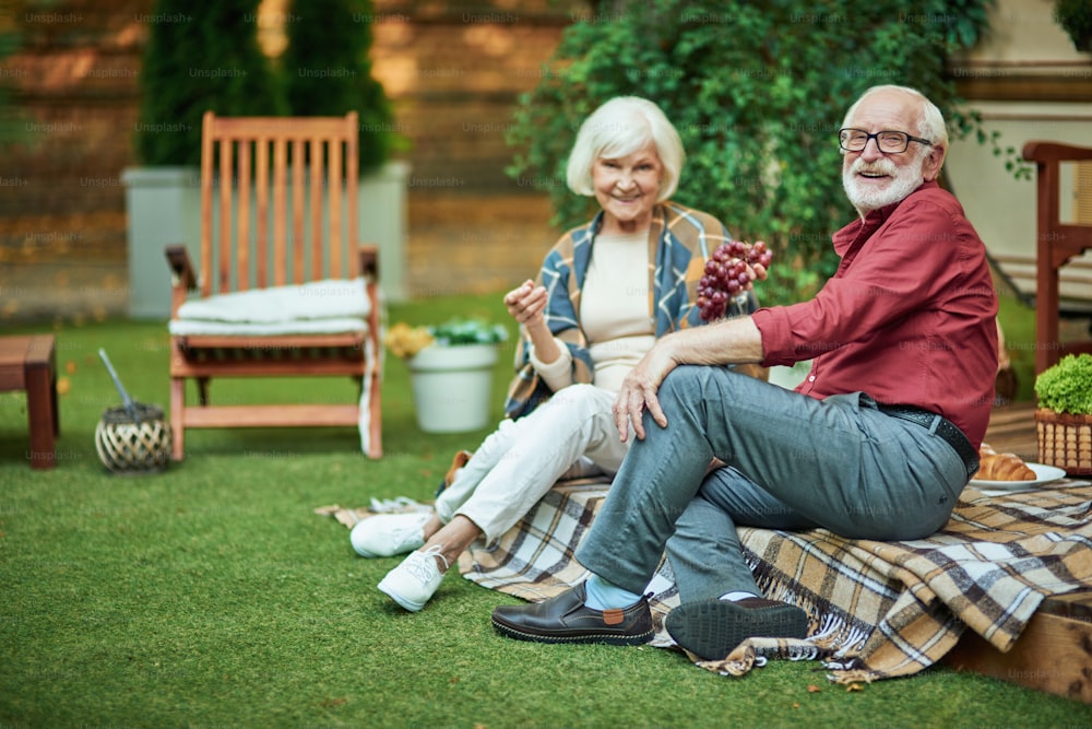 Full length photo of smiling senior couple sitting together on the van porch and eating grapes. Copy space. Relationship and family concept