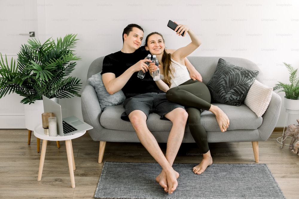 Attractive young sports yoga people, holding bottles of water, talking and smiling while resting after workout on gray sofa at home and making selfie photo on smartphone.