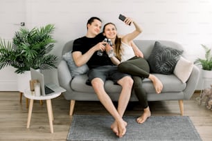 Attractive young sports yoga people, holding bottles of water, talking and smiling while resting after workout on gray sofa at home and making selfie photo on smartphone.