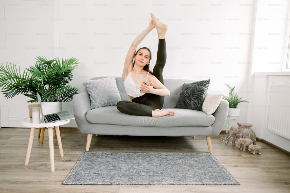 Pretty young Caucasian yoga fit woman sitting on gray sofa and posing to camera, stretching leg up, smiling and holding her smartphone. Woman watching online video yoga lessons.