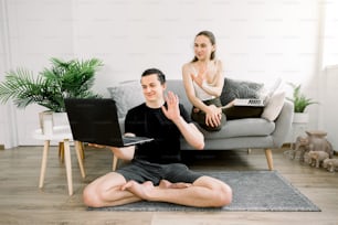 Young joyful couple of yoga coaches, sitting in lotus position, having fun at home after yoga workout, having video call on laptop with their friends, waving and smiling. Healthy lifestyle.