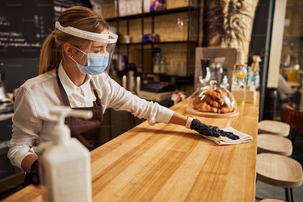 Side view portrait of beautiful woman in protective mask and gloves wiping wooden table with cloth