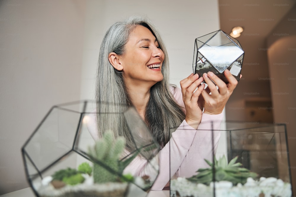 Senhora encantadora olhando para o recipiente de vidro geométrico da planta e sorrindo enquanto passa o tempo em casa