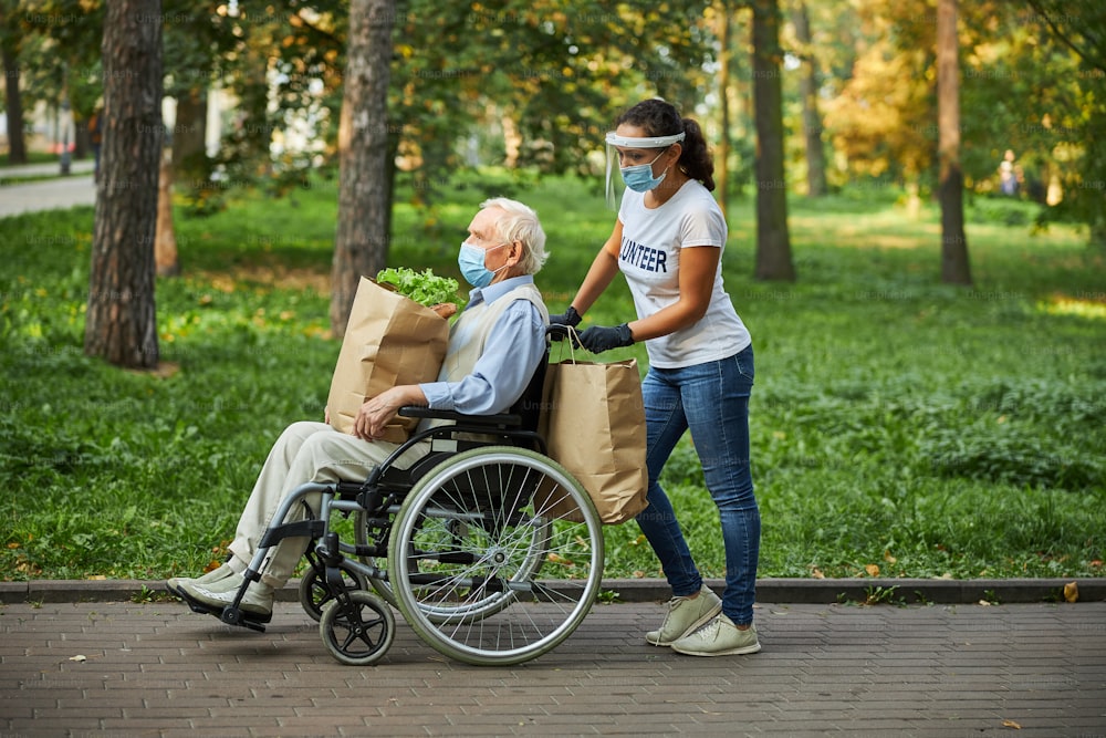 Full length side view portrait of friendly woman volunteer walking with elderly man in the city park or wood