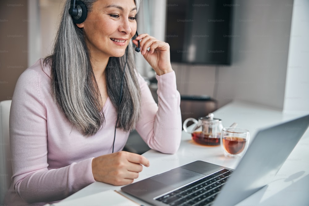 Smiling lady wearing headset while sitting at the table with notebook and talking with colleague through video call