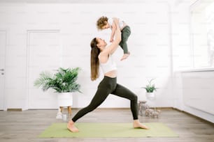 Cute little son and pretty young mother doing sport and yoga exercises in cozy yoga studio or home. Attractive sport woman stands on green yoga mat and raising up her little son.