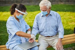 Female nurse using hand while screening vital sign of old patient while sitting on bench and wearing protection