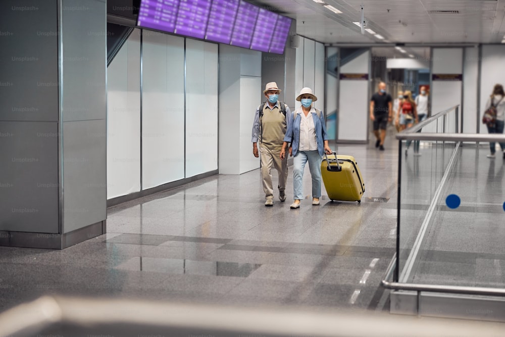 Ritratto a figura intera di due passeggeri anziani in maschere protettive usa e getta che percorrono il terminal dell'aeroporto