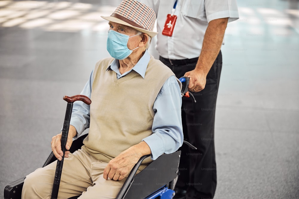 Thoughtful aged Caucasian male passenger in a disposable protective mask sitting in a transport chair