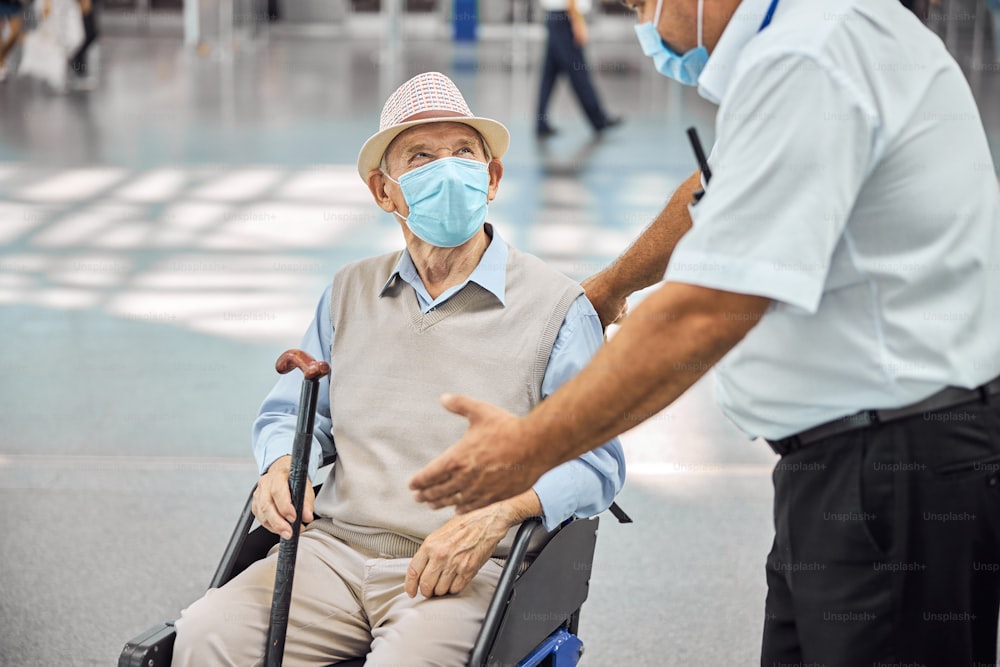 Caucasian airline male employee in a protective mask explaining something to a disabled elderly man