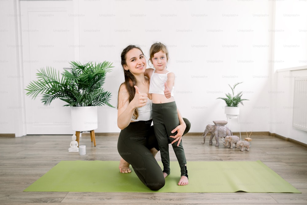 Family sport and yoga concept. Promotion of healthy lifestyle. Mother and little daughter smiling together at camera and showing thumbs up, while standing in cozy light yoga studio.