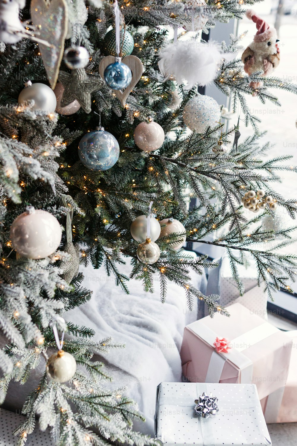 Stylish christmas tree with silver baubles, golden lights and wrapped presents in festive room. Modern decorated christmas tree with white balls, silver stars and toys. Merry Christmas