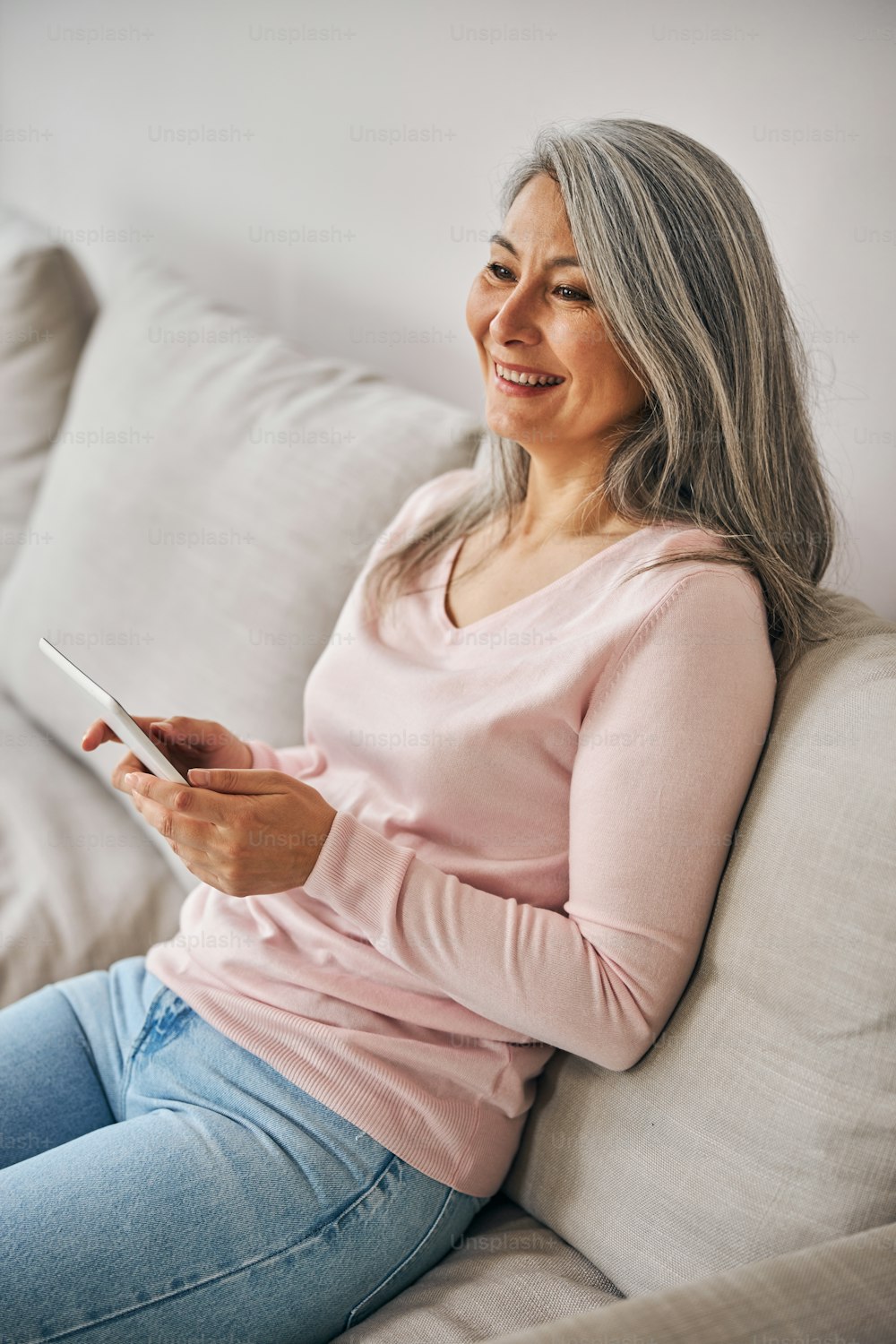 Side view portrait of laughing woman in pink sweater with modern tablet in hands sitting on the gray sofa in room indoors