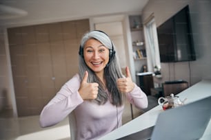 Beautiful happy lady in headphones with microphone showing approval gesture and smiling while sitting at the table with notebook