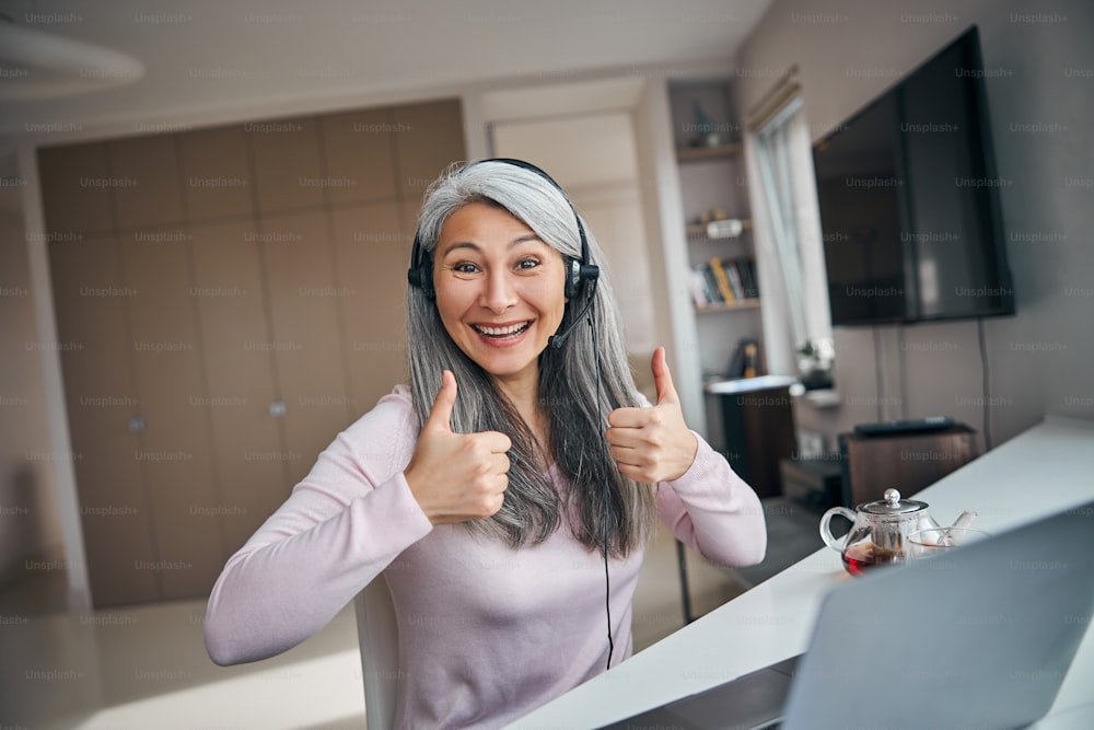 Beautiful happy lady in headphones with microphone showing approval gesture and smiling while sitting at the table with notebook