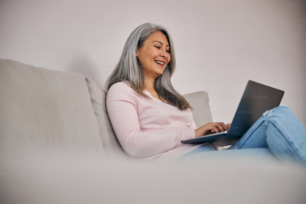 Close up portrait of happy smiling female entrepreneur looking to the screen modern technology device while sitting on the gray sofa in room indoors