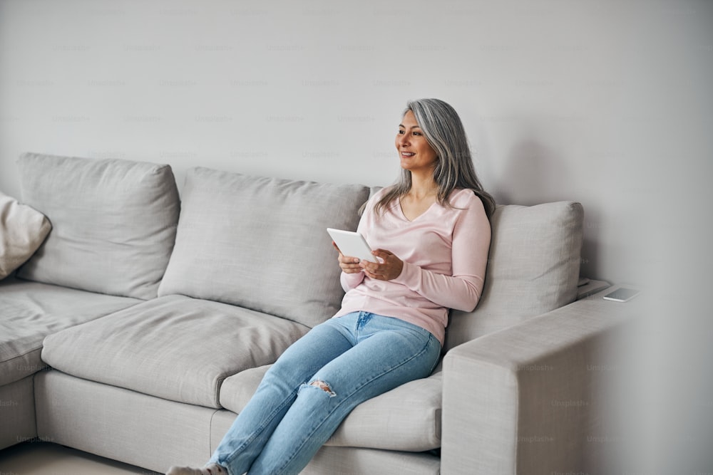 Full length portrait of happy smiling woman using modern tablet while sitting on gray sofa in living room