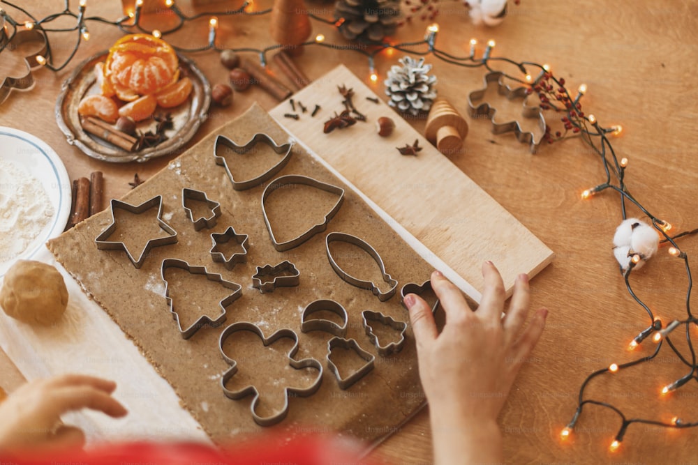 Person making Christmas gingerbread cookies, holiday advent. Hands cutting gingerbread dough with festive metal cutters on rustic table with spices, oranges, festive decorations, lights