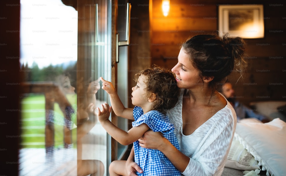 Small girl with parents by window in wooden cabin, holiday in nature concept.