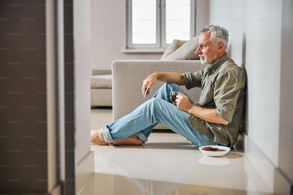 Uomo anziano calmo con una tazza di tè e uno smartphone seduto sul pavimento vicino a un barattolo di biscotti