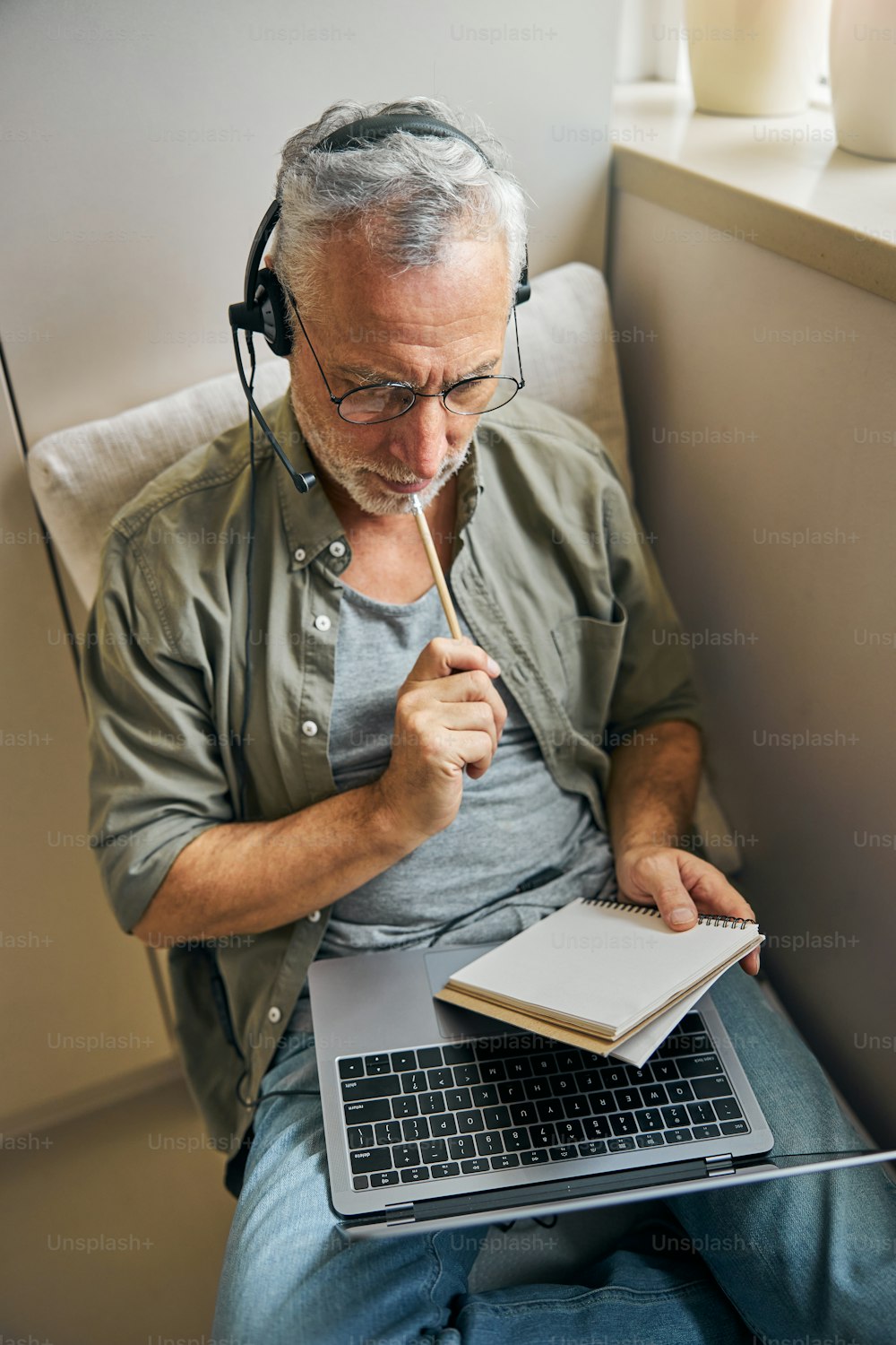 Anciano inteligente con una computadora portátil que se ve pensativo mientras se toca los labios con un lápiz y usa auriculares