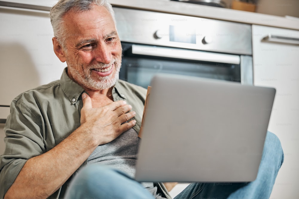 Close-up photo of a grey-haired man touching his chest and looking gratefully at a computer screen