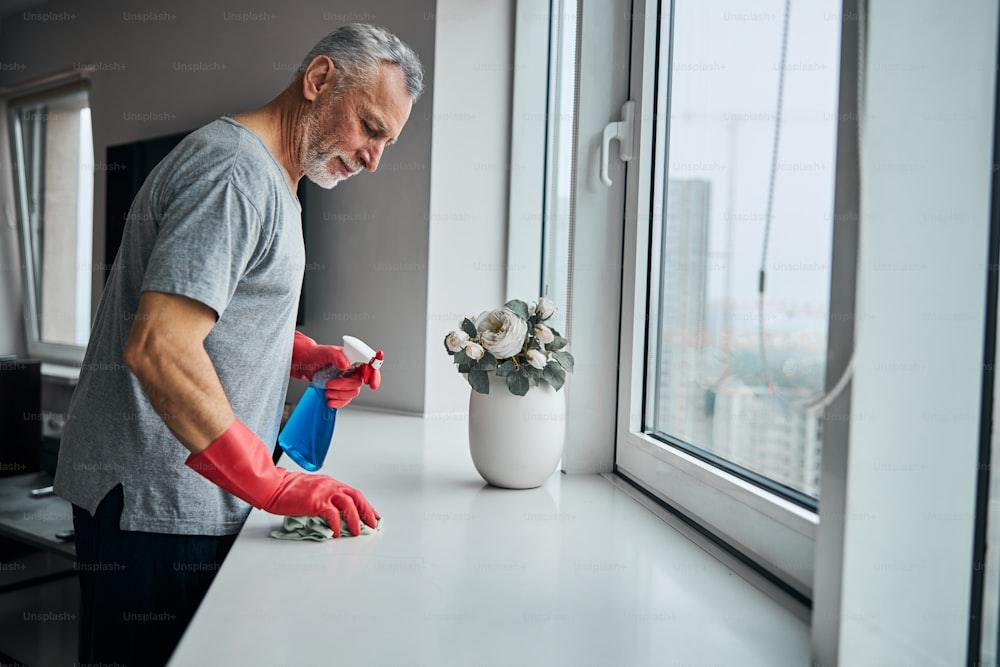 Careful elderly gentleman in rubber gloves cleaning a windowsill in his apartment by wiping off the dust