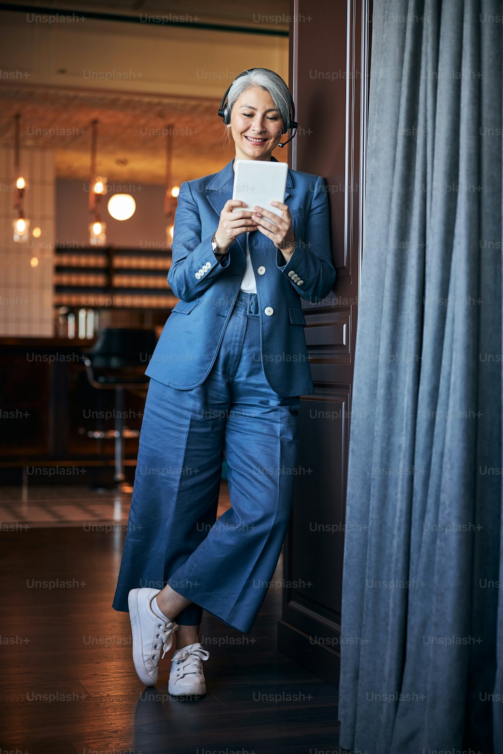 Schöne Dame im eleganten Anzug, die einen Tablet-Computer hält und lächelt, während sie sich an die Tür lehnt