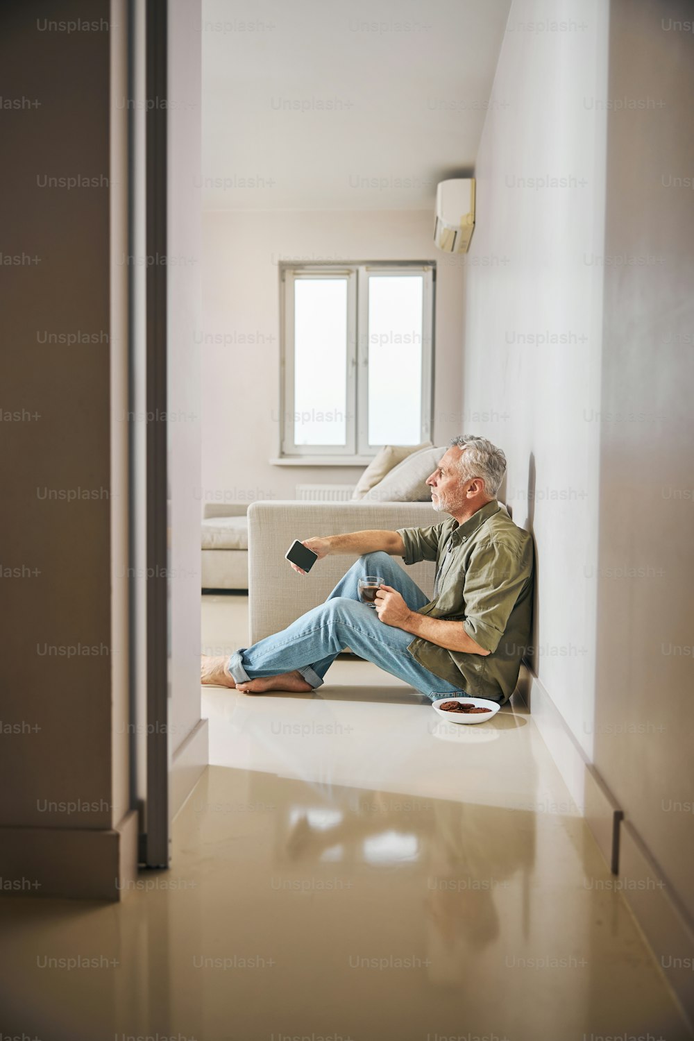 Uomo anziano vestito in modo casual seduto sul pavimento con una tazza di tè e il suo smartphone