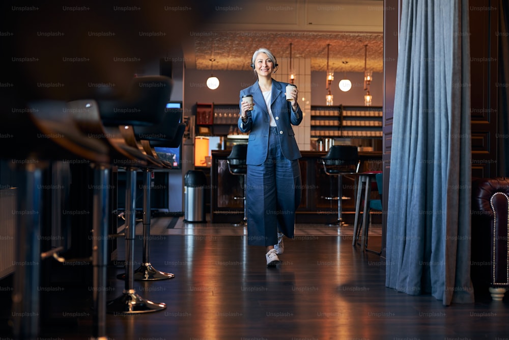 Charming lady in elegant suit holding two takeout coffee cups and smiling while strolling down the cafeteria
