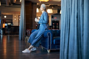 Elegante dama con traje azul mirando hacia otro lado y sonriendo mientras sostiene una taza de café para llevar