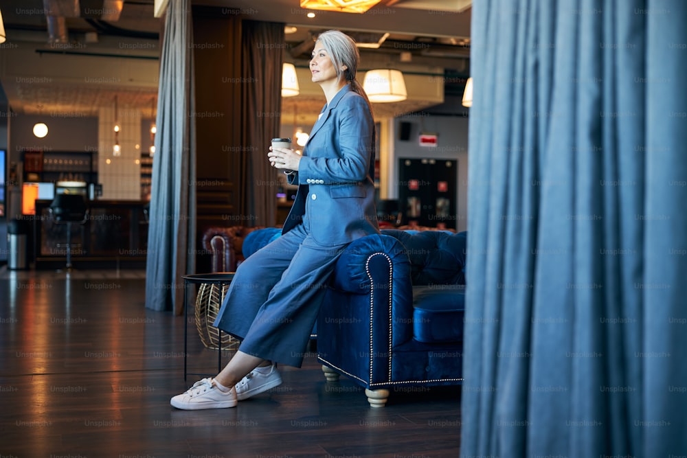 Elegant lady in blue suit looking away and smiling while holding takeaway cup of coffee