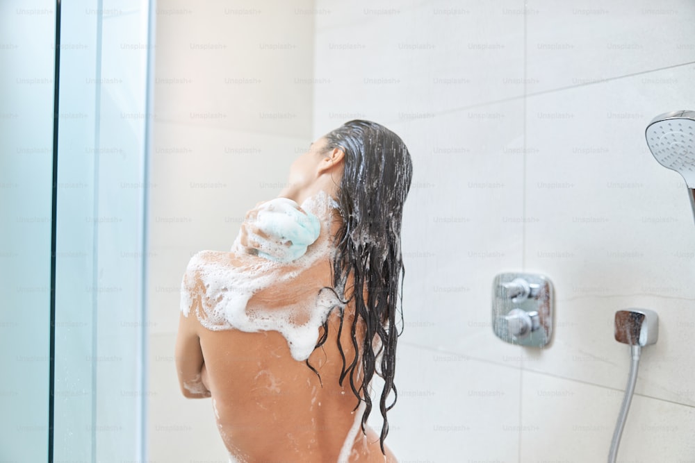Vista posteriore della signora nuda con lunghi capelli bagnati usando una spugna da bagno esfoliante mentre fa la doccia a casa