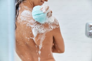 집에서 샤워를 하는 동안 각질 제거 목욕 스폰지를 사용하여 피부에 거품이 있는 여성을 닫습니다.